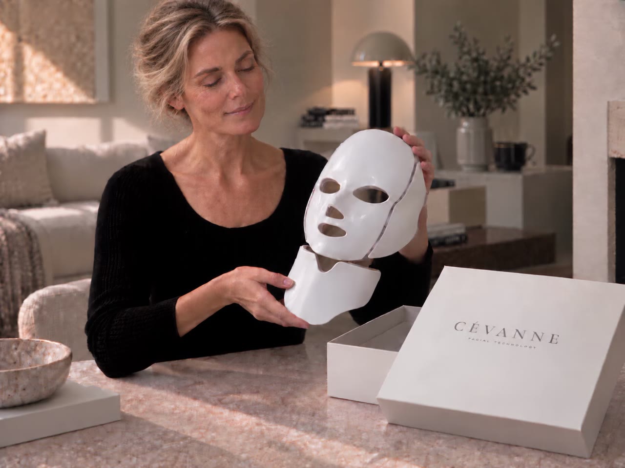 A woman holds the CÉVANNE mask and switches it on; the red light rises softly inside the shell.